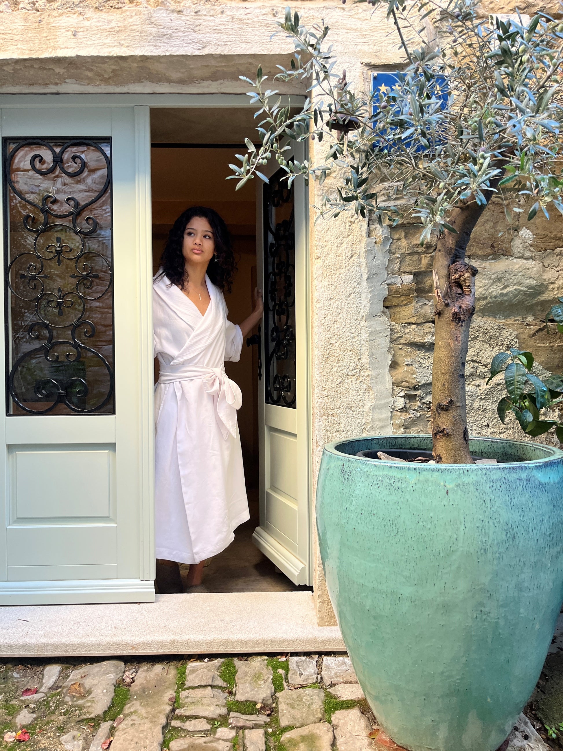 An image of a beautiful lady in a white linen robe opening the front door to an old stone home.