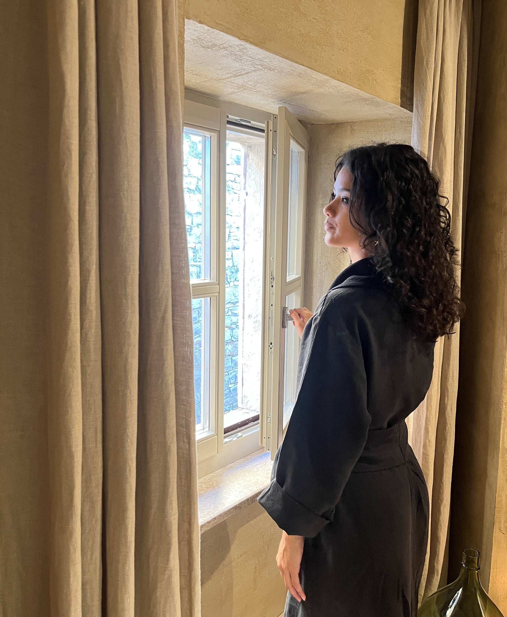 Side view of a woman in a black linen robe opening a window in the morning.