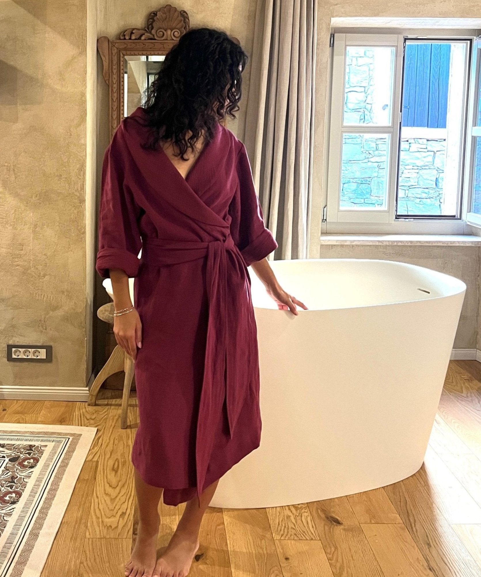 A woman in a luxurious burgundy robe, standing beside a tub and looking to the side.