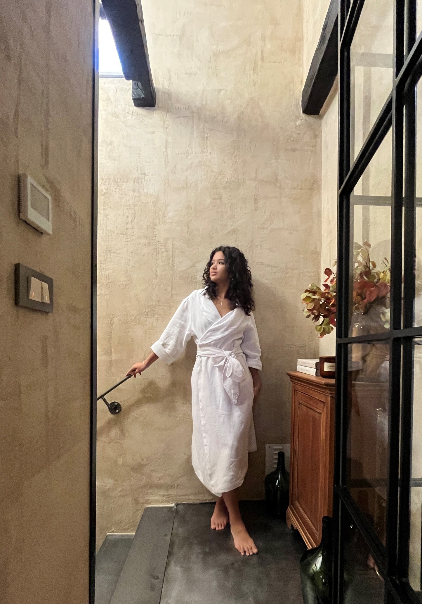 Woman in a white linen robe standing in a stairway with a skylight.